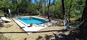 A rectangular pool with a clear blue water, surrounded by a gravel area and shaded by trees. There are lounge chairs and a hammock near the pool.