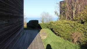 Un pont de fusta que condueix a una vista del mar, envoltat de vegetació i un edifici amb façana vertical.
