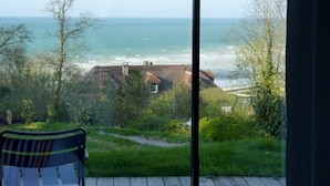 Vista d'una habitació amb una finestra gran que dona a una platja, un camí i vegetació.