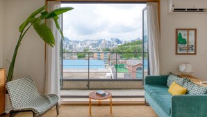Ein Zimmer mit großem Fenster, einem grünen Sofa, einem gemusterten Sessel und einem kleinen Tisch mit einem Buch.