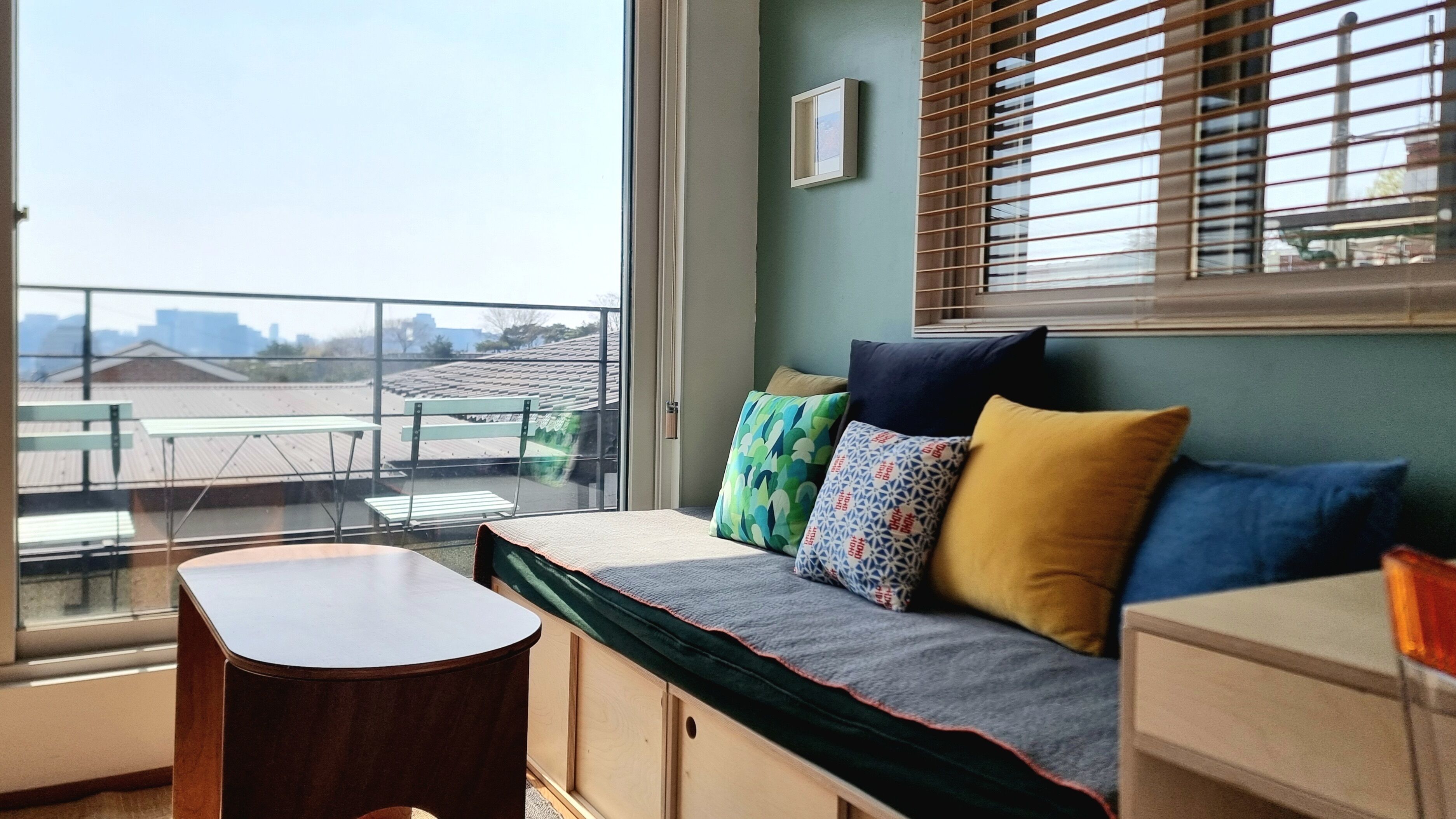 Huone, josta avautuu näkymä kaupungin siluettiin suurten ikkunoiden kautta, värikkäillä tyynyillä varustettu puinen penkki ja pieni puinen pöytä.