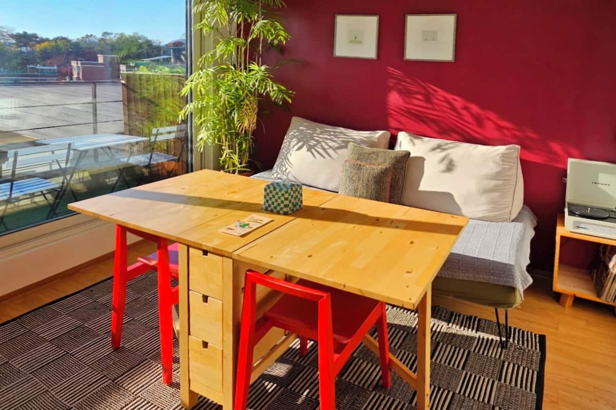 A room with a wooden table, red chairs, a sofa with cushions, a laptop on a side table, and a large window with a view of the outside.