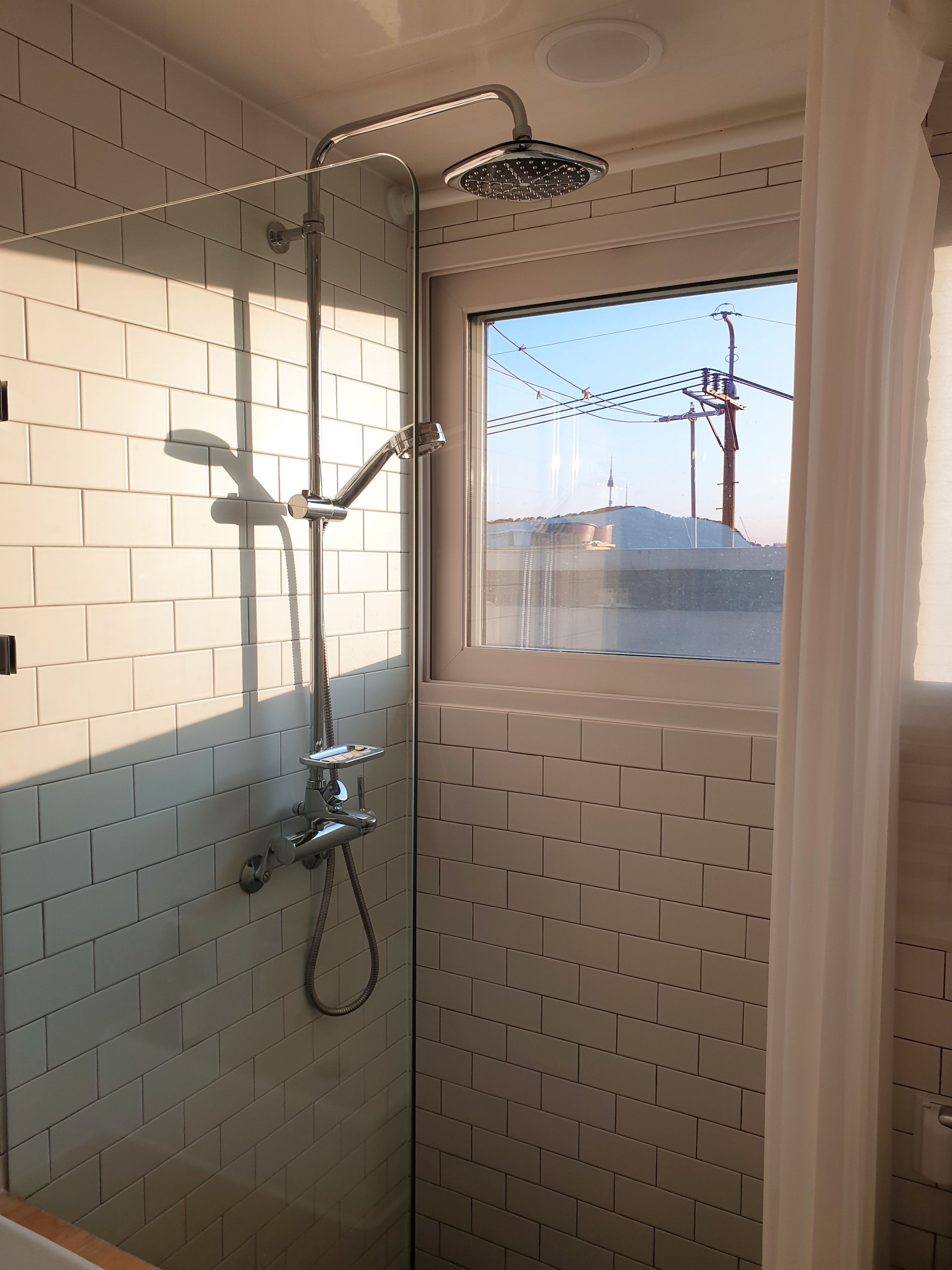 A shower with a glass door, white tiled walls, and a window overlooking a cityscape.