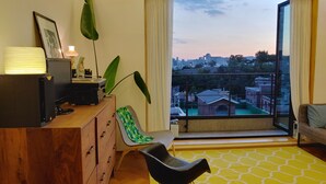 Ein modernes Wohnzimmer mit einem Holztisch, einem schwarzen Sessel, einem großen Fenster mit Blick auf die Stadt und einem gelb gemusterten Teppich.