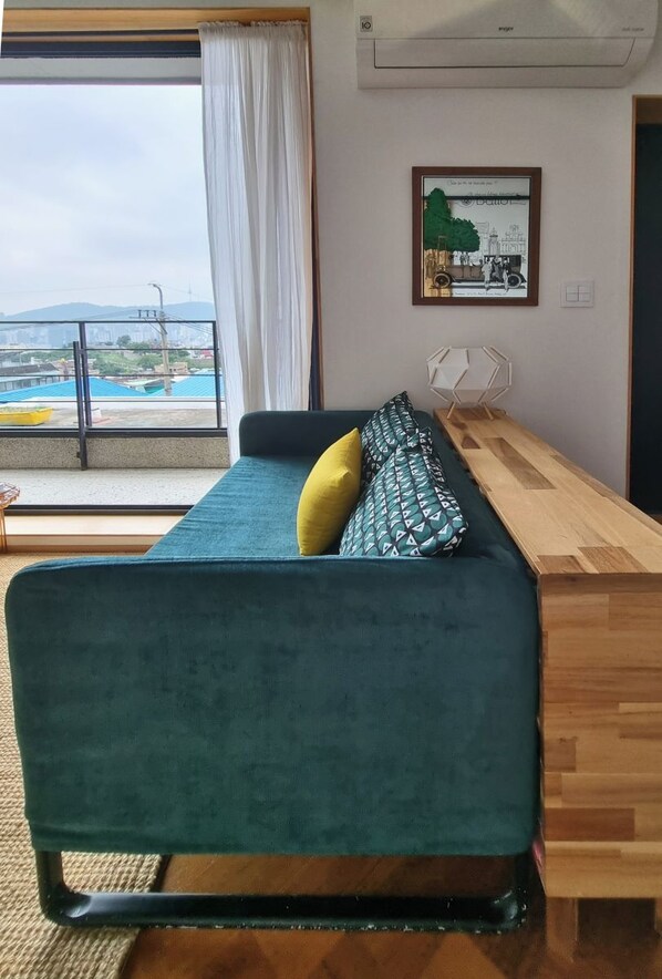 A room with a sofa, a wooden side table, a framed picture on the wall, and a view through a window. - Naksan Balcony (Seoul)
