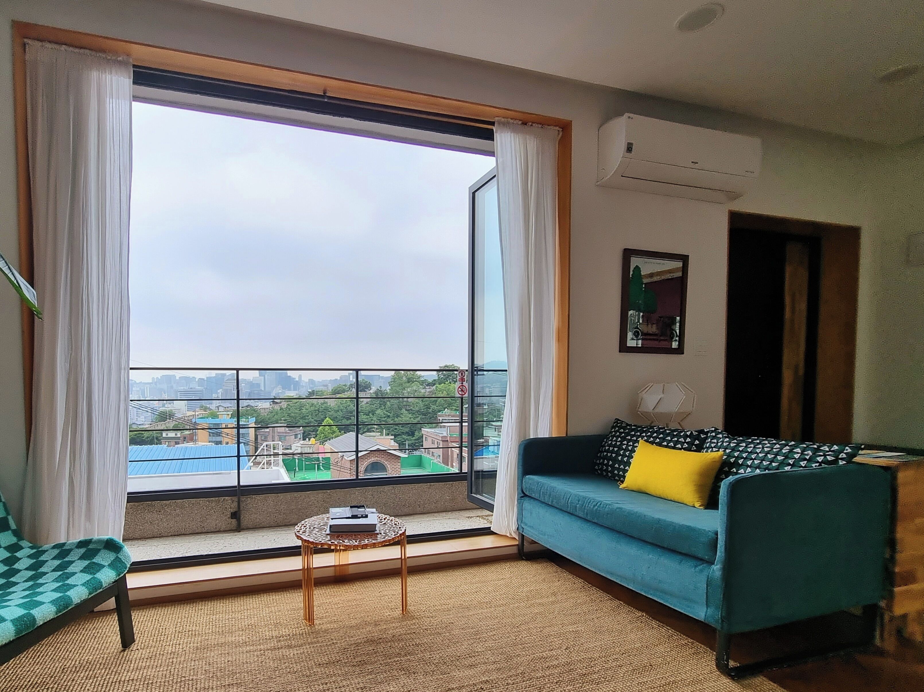 A living room with a large window, a blue sofa with a yellow pillow, a small round table, and a city view.