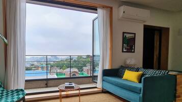 A living room with a large window, a blue sofa with a yellow pillow, a small round table, and a city view.