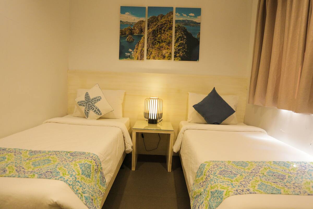 A hotel room with two beds, a nightstand with a lamp, and a wall with three framed pictures.
