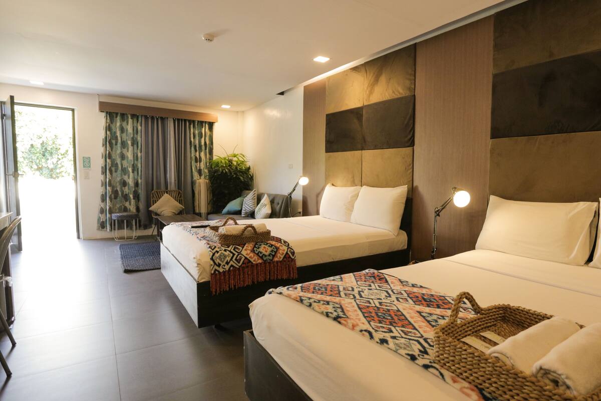 A hotel room with two beds, a large window with curtains, a plant, and a desk with a chair.