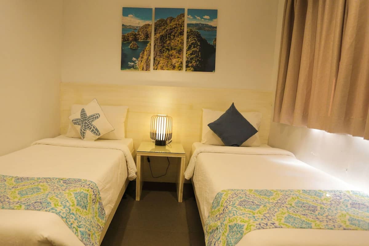 A hotel room with two beds, a nightstand with a lamp, a window with curtains, and a wall with three framed pictures.
