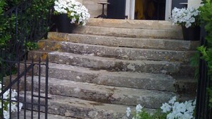 Una escalera de piedra que lleva a una puerta con contraventanas, rodeada de flores blancas en macetas y un portón de metal.