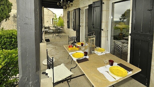 Un área de comedor al aire libre con una larga mesa preparada para dos, rodeada de muros de piedra y contraventanas negras.