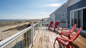 Une terrasse en bois avec des chaises rouges, une balustrade blanche et une vue sur la plage et l’océan.