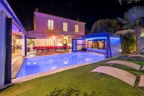 A swimming pool with a covered area, a patio with red chairs, and a house in the background.