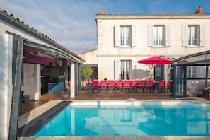 A swimming pool with red chairs and umbrellas, a wooden deck, and a building with windows and shutters in the background. - Villa-4 étoiles-Oversized-Whirlpool bath-Terrace (Angoulins)