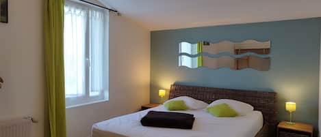 Un dormitorio con una cama grande, cortinas verdes, cabecera de mimbre, dos lámparas de noche y un espejo fijado a la pared.