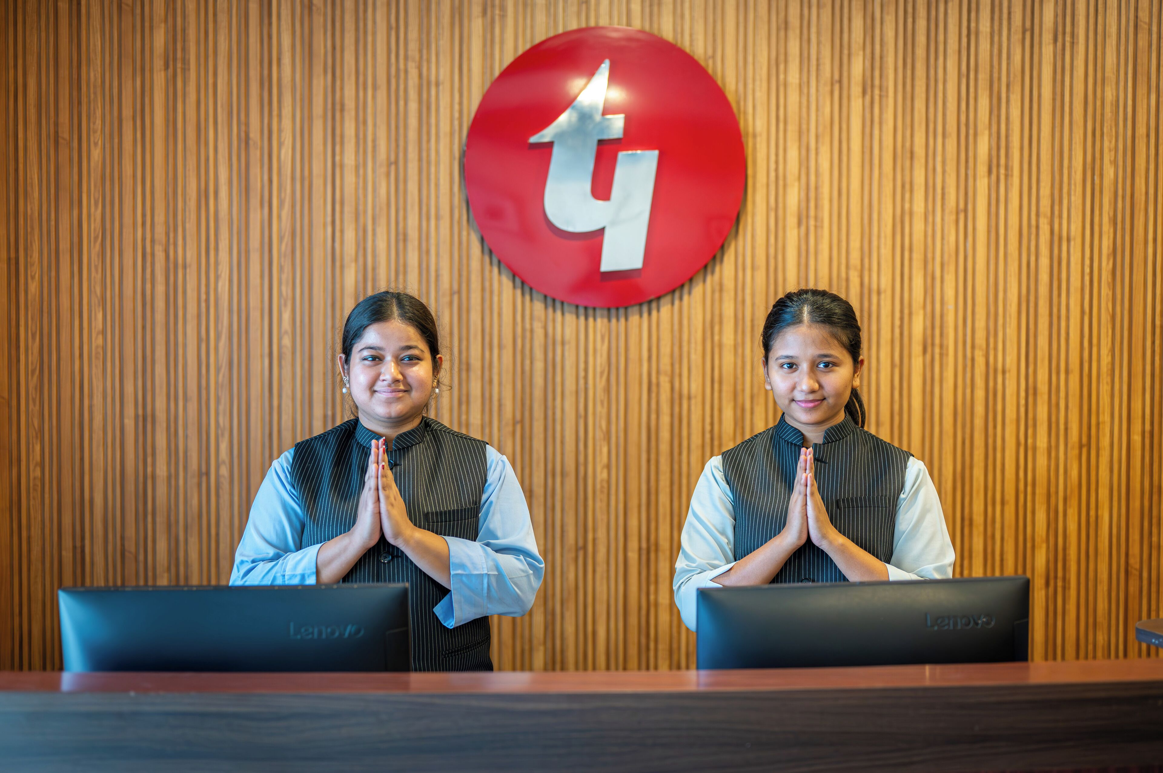 Two individuals seated at desks with laptops, in front of a wooden wall with a red circular logo.