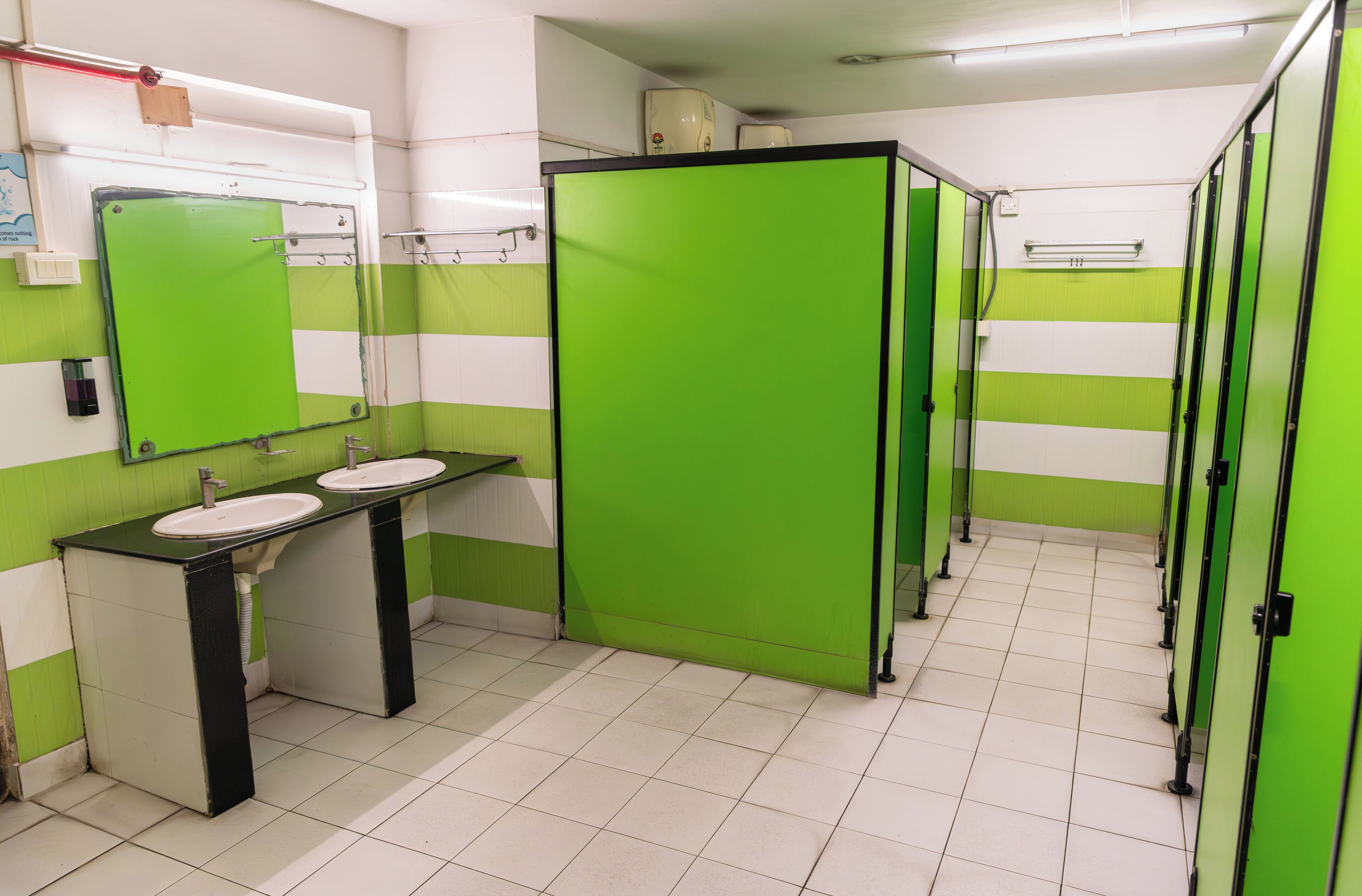 Baño con separadores verdes, pisos de baldosas blancas y dos lavamanos con encimeras negras.