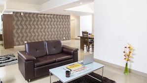 A modern living room with a brown leather sofa, a coffee table with a laptop and magazines, a dining area with a wooden table and chairs, and a patterned wallpaper wall. - SKYLA Serviced Apartments Banjara Hills (Hyderabad)