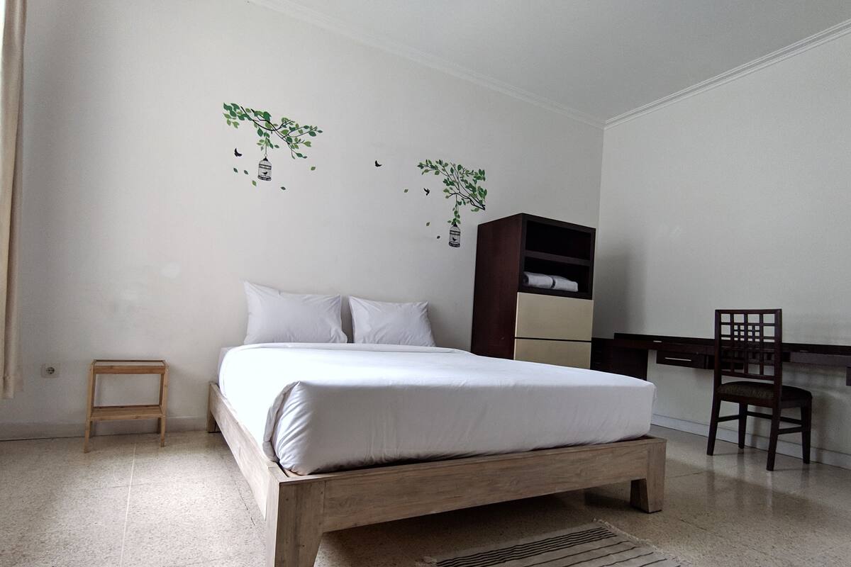 Un dormitorio con una cama, un taburete de madera, un escritorio con silla y un armario.
