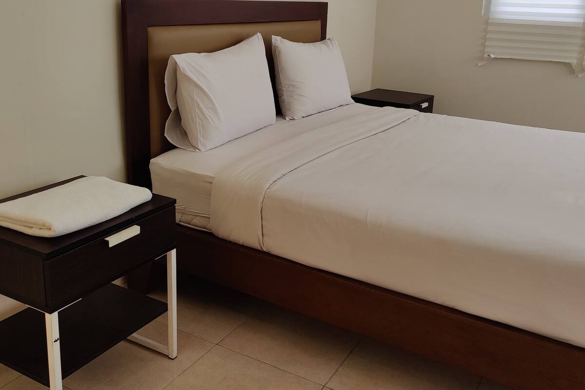 Una cama bien tendida con ropa blanca, un cabecero de madera y una mesita de noche con una toalla blanca.