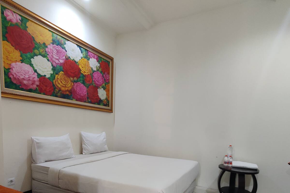 Un dormitorio ordenado con una cama, una mesita con botellas y un cuadro floral colorido en la pared.