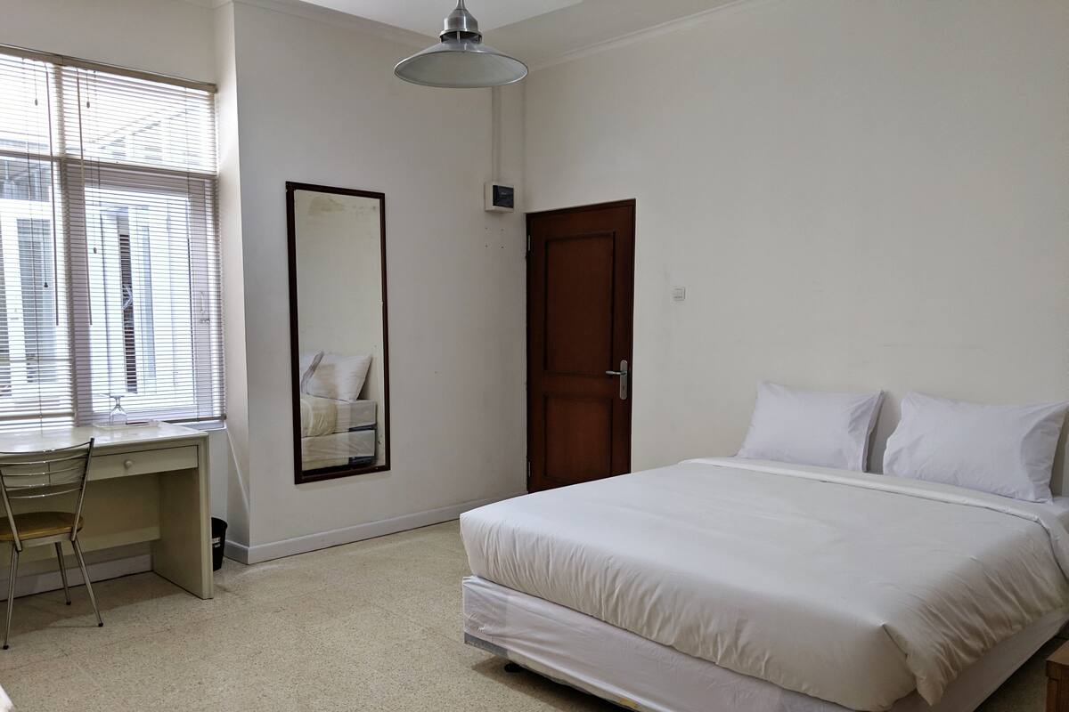 Un dormitorio con una cama, un escritorio, una silla, un espejo grande y una puerta.
