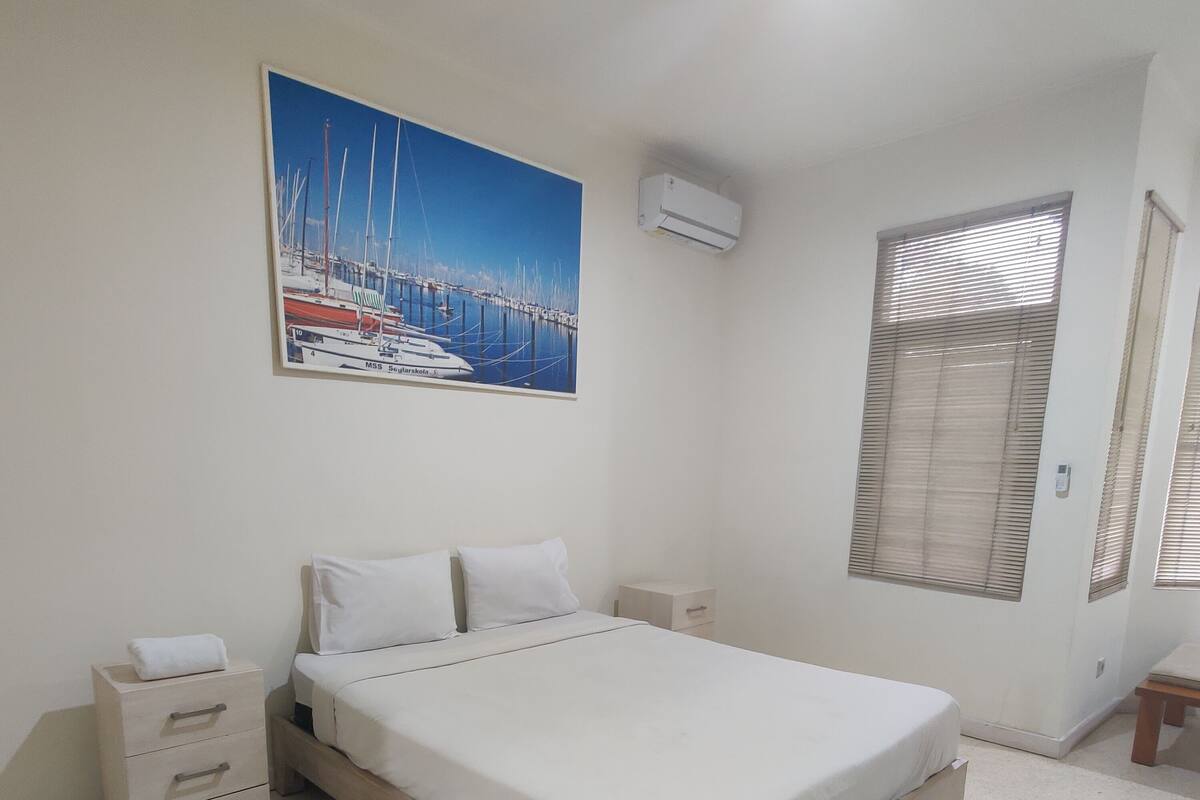 Un dormitorio con cama, mesita de noche, cómoda, un cuadro de un puerto deportivo y ventana con persianas.