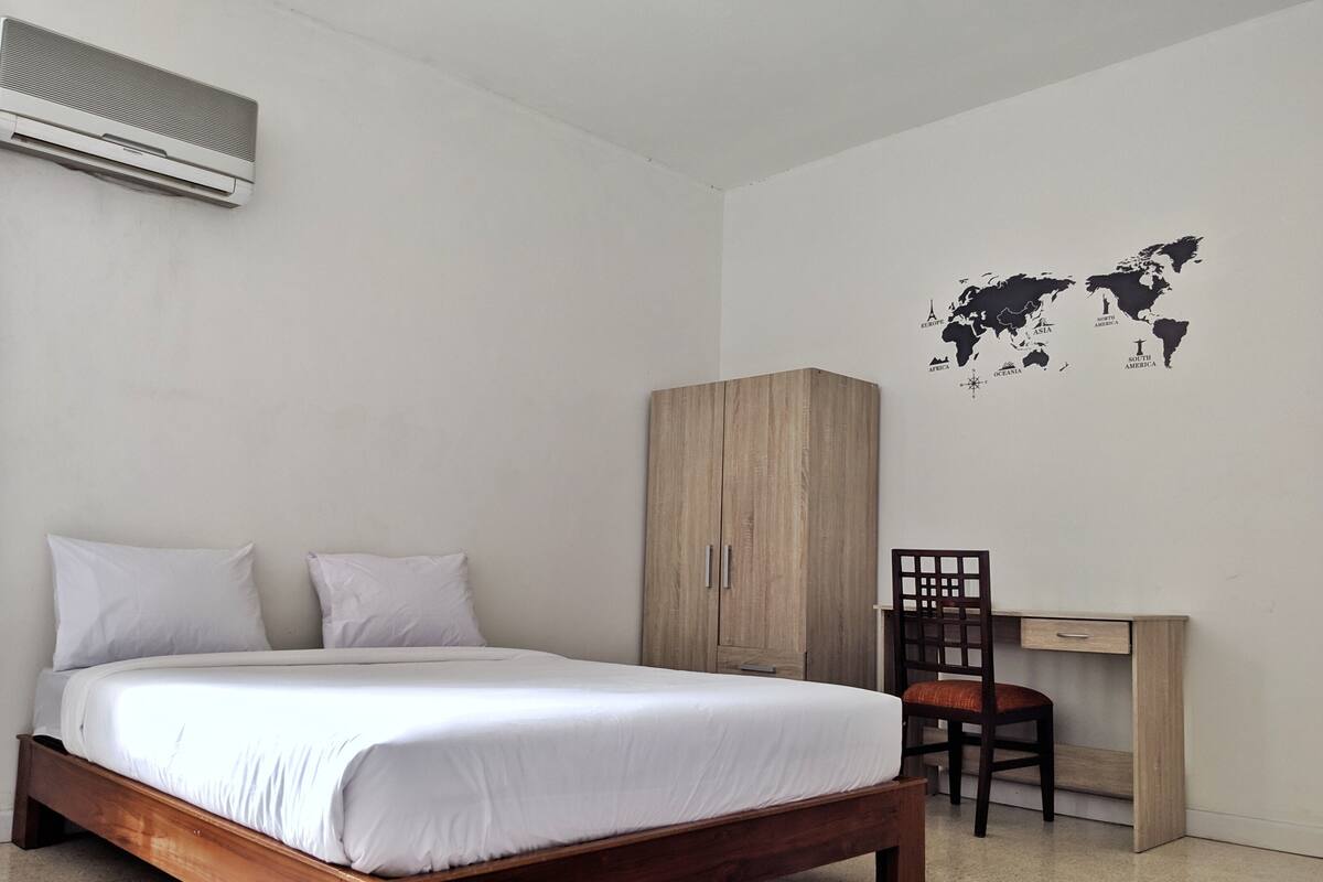 Un dormitorio con cama de madera, ropa de cama blanca, un armario, un escritorio con silla y un vinilo decorativo de un mapa del mundo.