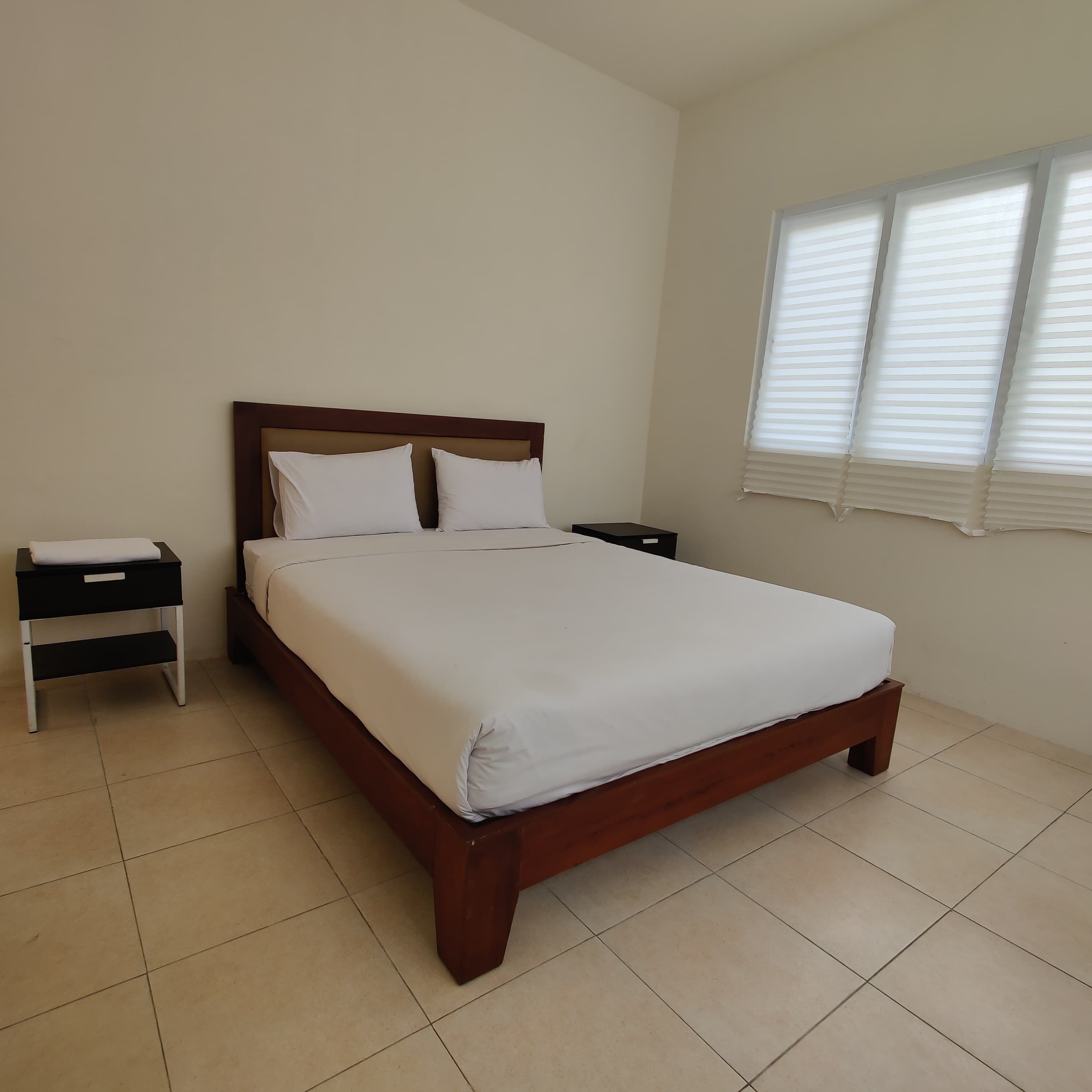 Un dormitorio con cama de madera, ropa de cama blanca, una mesita de noche y una ventana con persianas.