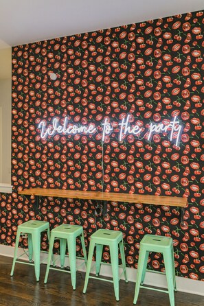 Un mur avec un fond noir et un motif de cerises rouges. Un panneau néon affiche l’inscription « Bienvenue à la fête ». Trois tabourets verts sont placés devant le mur.