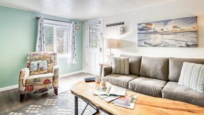 Een woonkamer met een gestreept fauteuil, een grijze bank, een houten salontafel en een schilderij van een strandgezicht.