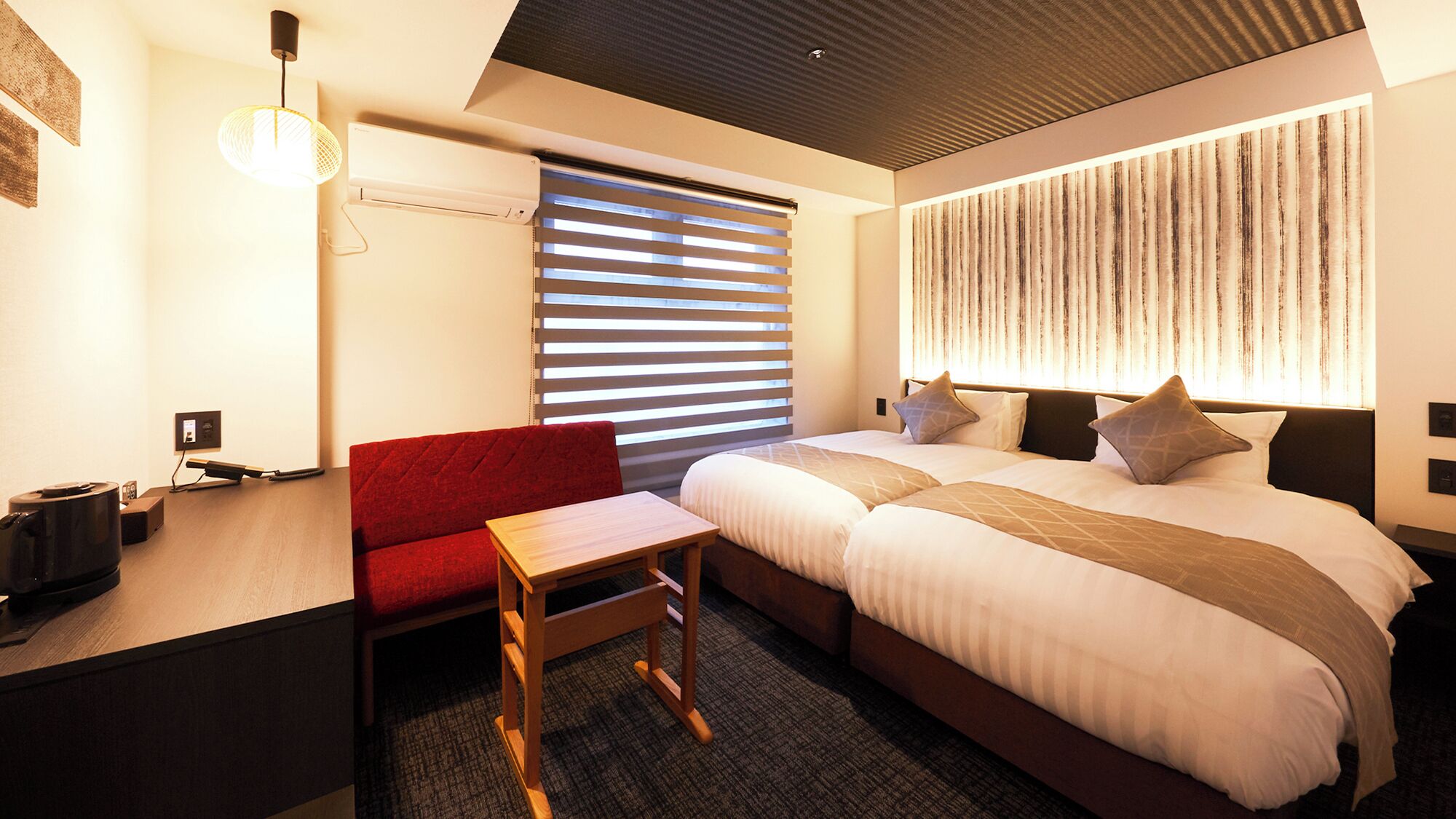 A hotel room with a bed, a desk, a chair, a small table, a wall-mounted air conditioner, and a window with blinds.