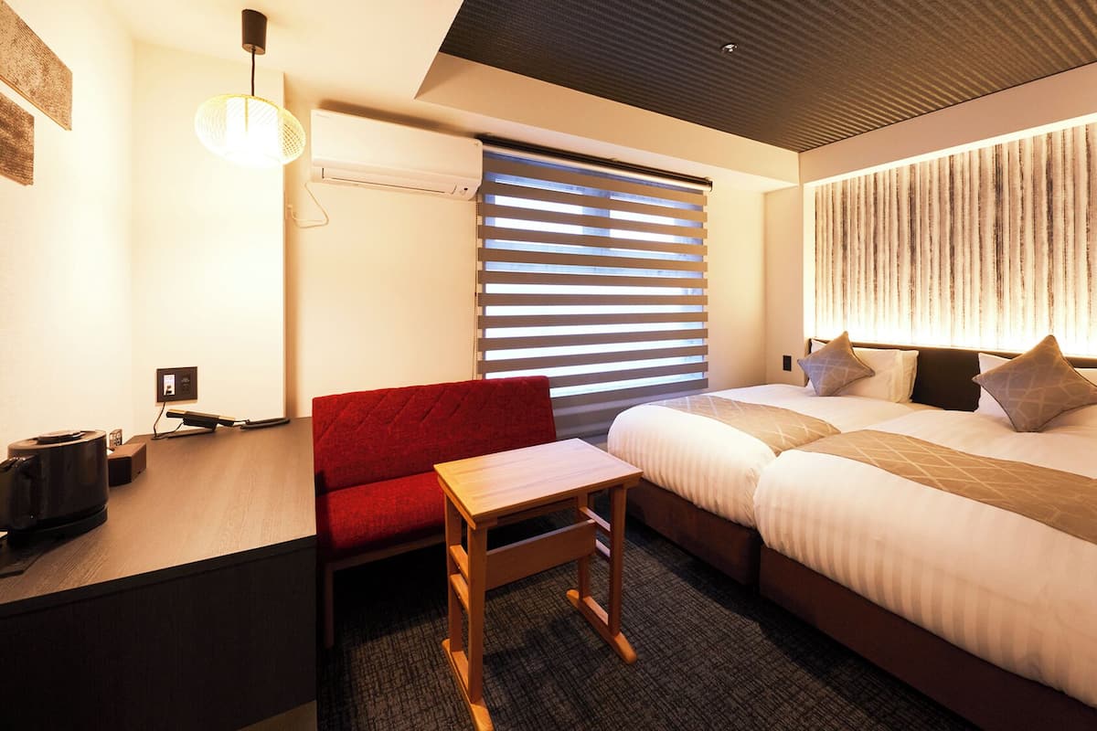 A hotel room with a bed, a desk, a chair, a small table, a wall-mounted air conditioner, and a window with blinds.