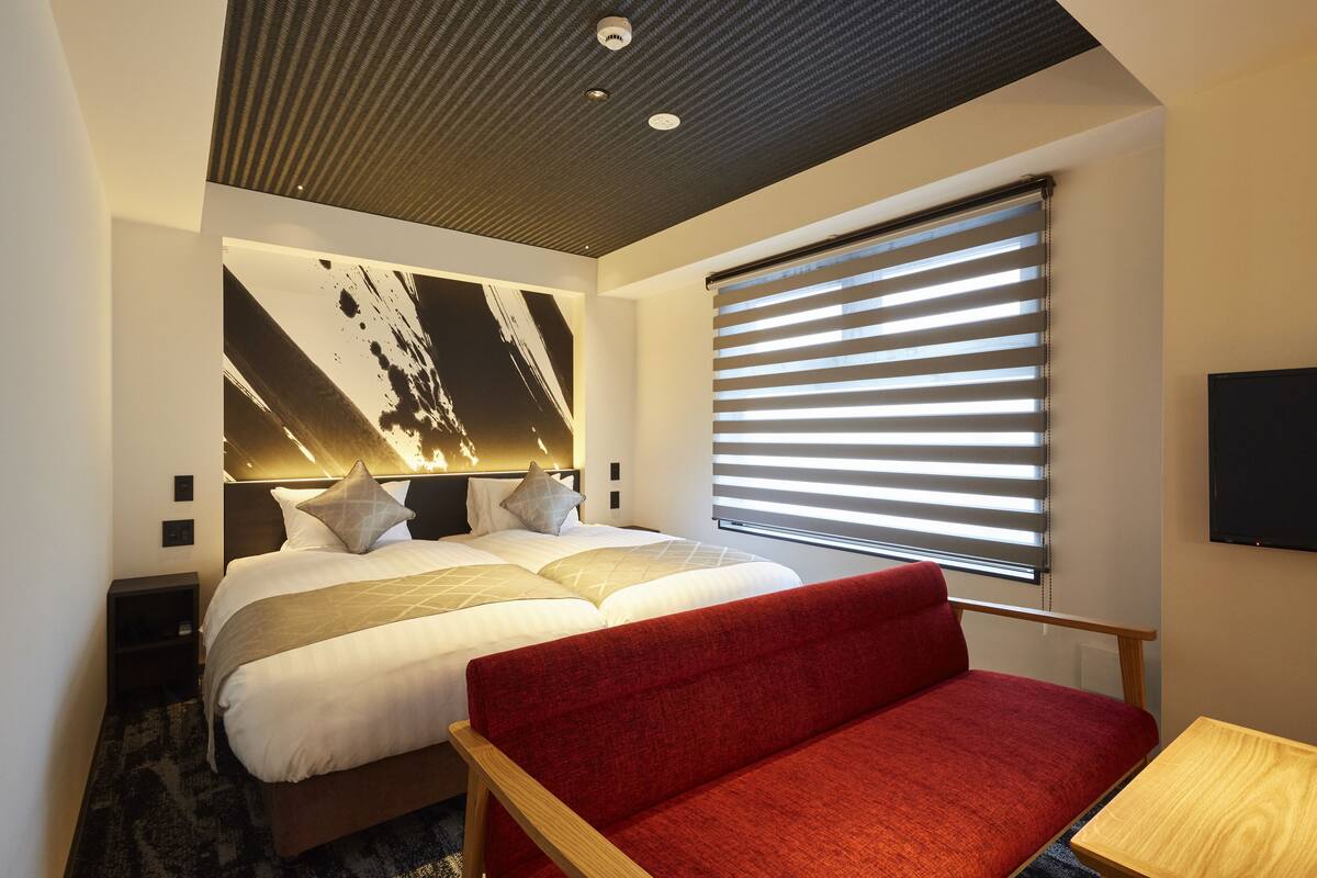 A modern hotel room with a large bed, a red sofa, a flat-screen TV, and a large abstract painting on the wall.