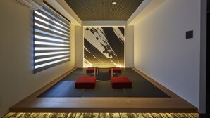 Une pièce moderne avec un plafond en pente, une grande œuvre d’art abstraite accrochée au mur, une petite table ronde avec quatre coussins rouges et une fenêtre avec des stores horizontaux.