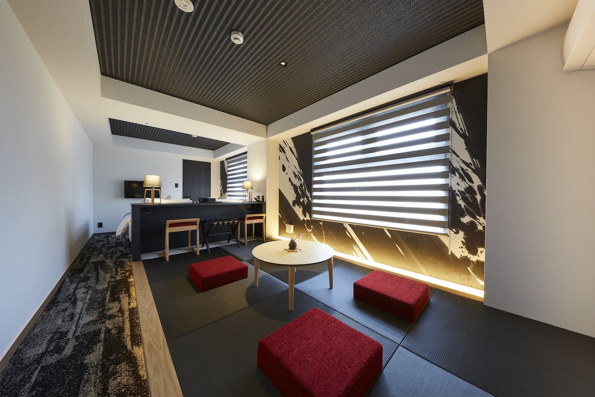 A modern interior with a kitchen area, seating with red cushions, and a large window with blinds.
