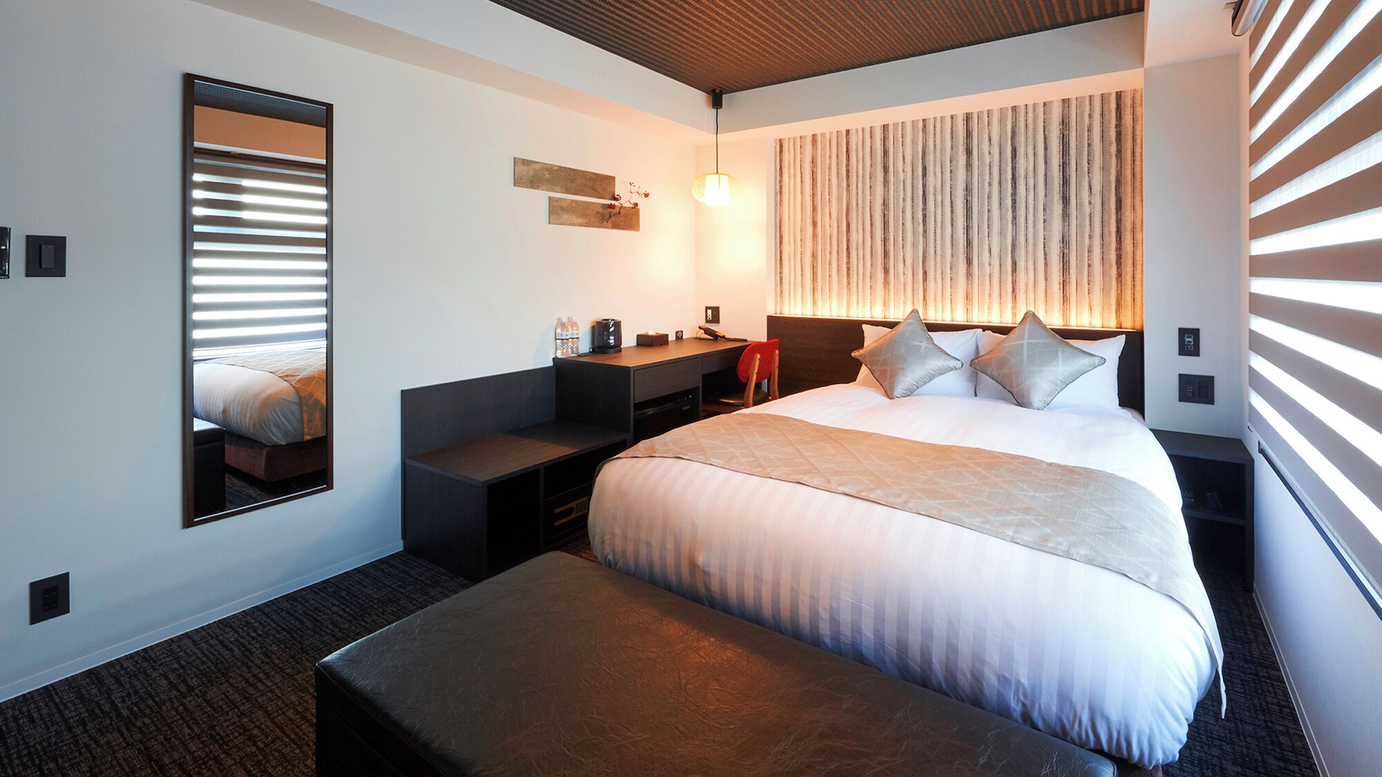 A hotel room with a bed, a desk, a chair, a mirror, and a wall-mounted light fixture.