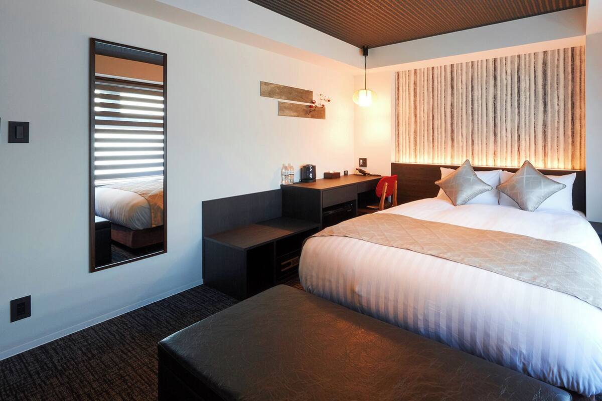 A hotel room with a bed, a desk, a chair, a mirror, and a wall-mounted light fixture.