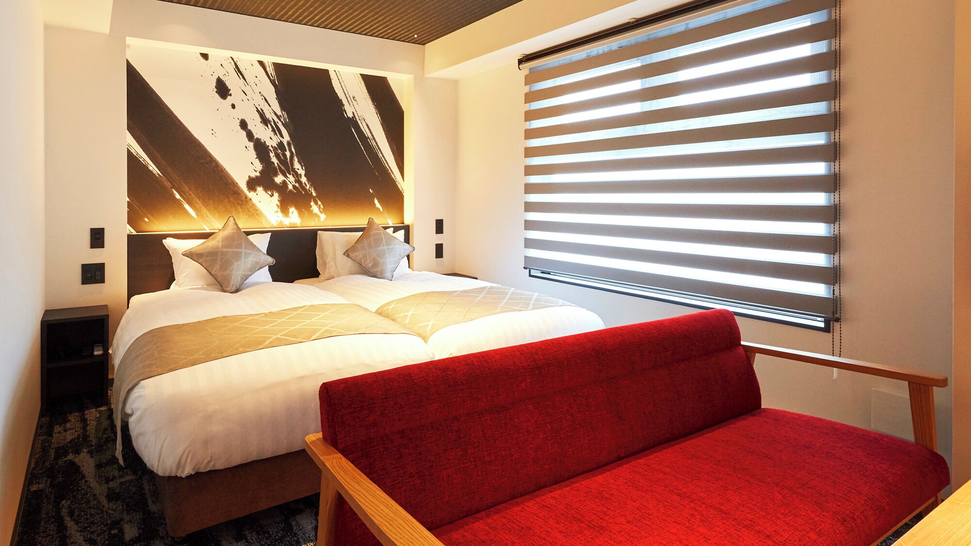 A hotel room with a large bed, a red sofa, a wall-mounted TV, and a window with blinds.