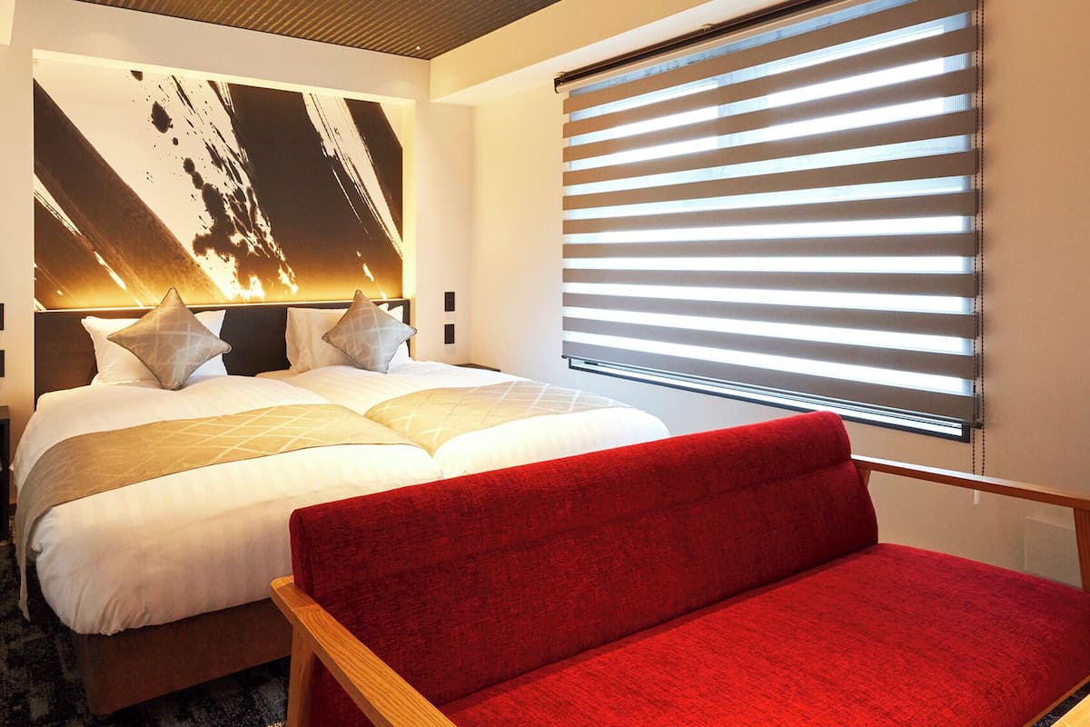 A hotel room with a large bed, a red sofa, a wall-mounted TV, and a window with blinds.