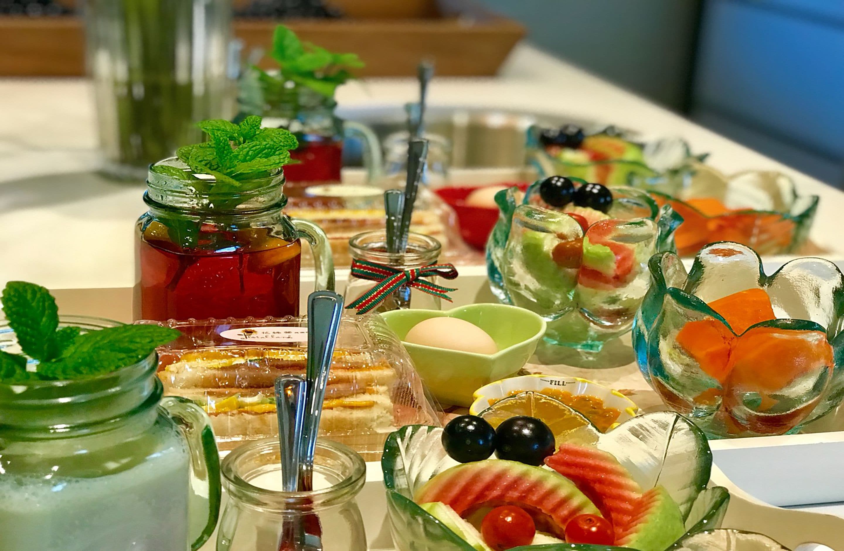 Variedad de alimentos y bebidas sobre una mesa, incluyendo frascos con bebidas, un bol de fruta y un pastel.