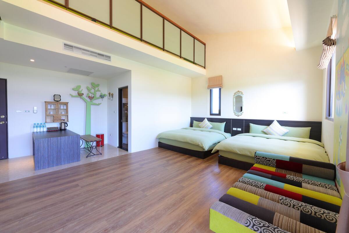 A modern hotel room with a wooden floor, two beds, a small desk with a chair, a decorative tree wall, and a large window.