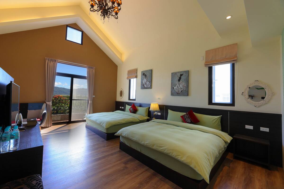 A bedroom with two beds, a desk with a chair, a large window, and artwork on the walls.