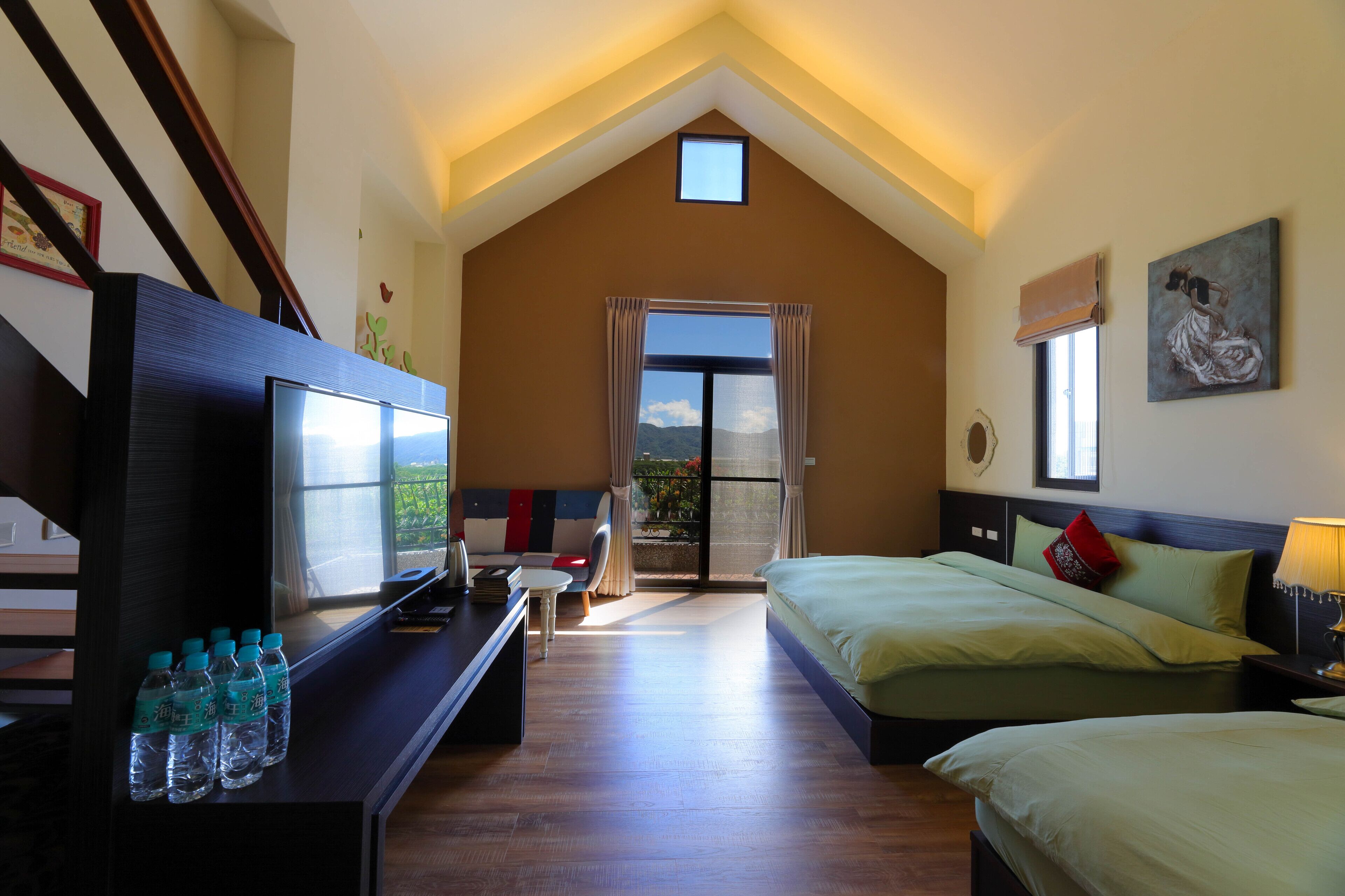 Ein Schlafzimmer mit Fernseher, Schreibtisch mit Wasserflaschen, Bett mit Kissen, Nachttisch und einem Fenster mit Vorhängen.