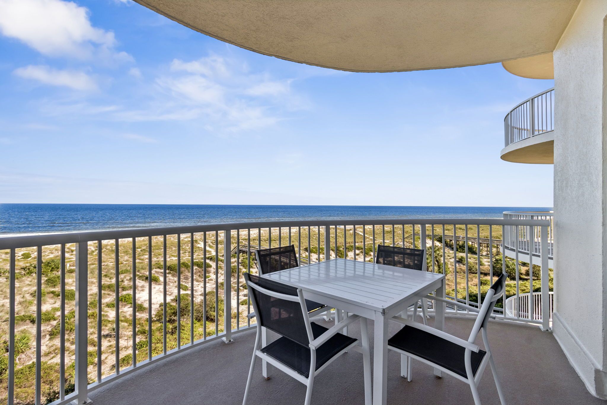 A balcony with a table and chairs, overlooking a beach with a clear sky.