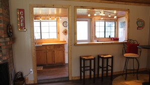 Un intérieur chaleureux avec un comptoir en bois, des tabourets, une cheminée et une cuisine visible à travers une porte ouverte.