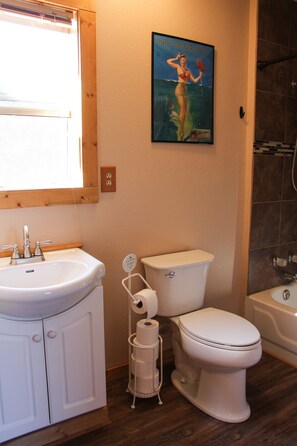 Ein Badezimmer mit einem weißen Waschbecken, einer Toilette und einer Badewanne. An der Wand hängt ein Poster und über dem Waschbecken befindet sich ein Fenster.