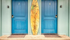 Duas portas azuis com os números 7 e 6, uma prancha de surf encostada às portas e um tapete de entrada à frente.