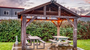Um gazebo de madeira com duas mesas de piquenique, cercado por vegetação e um sebedo.